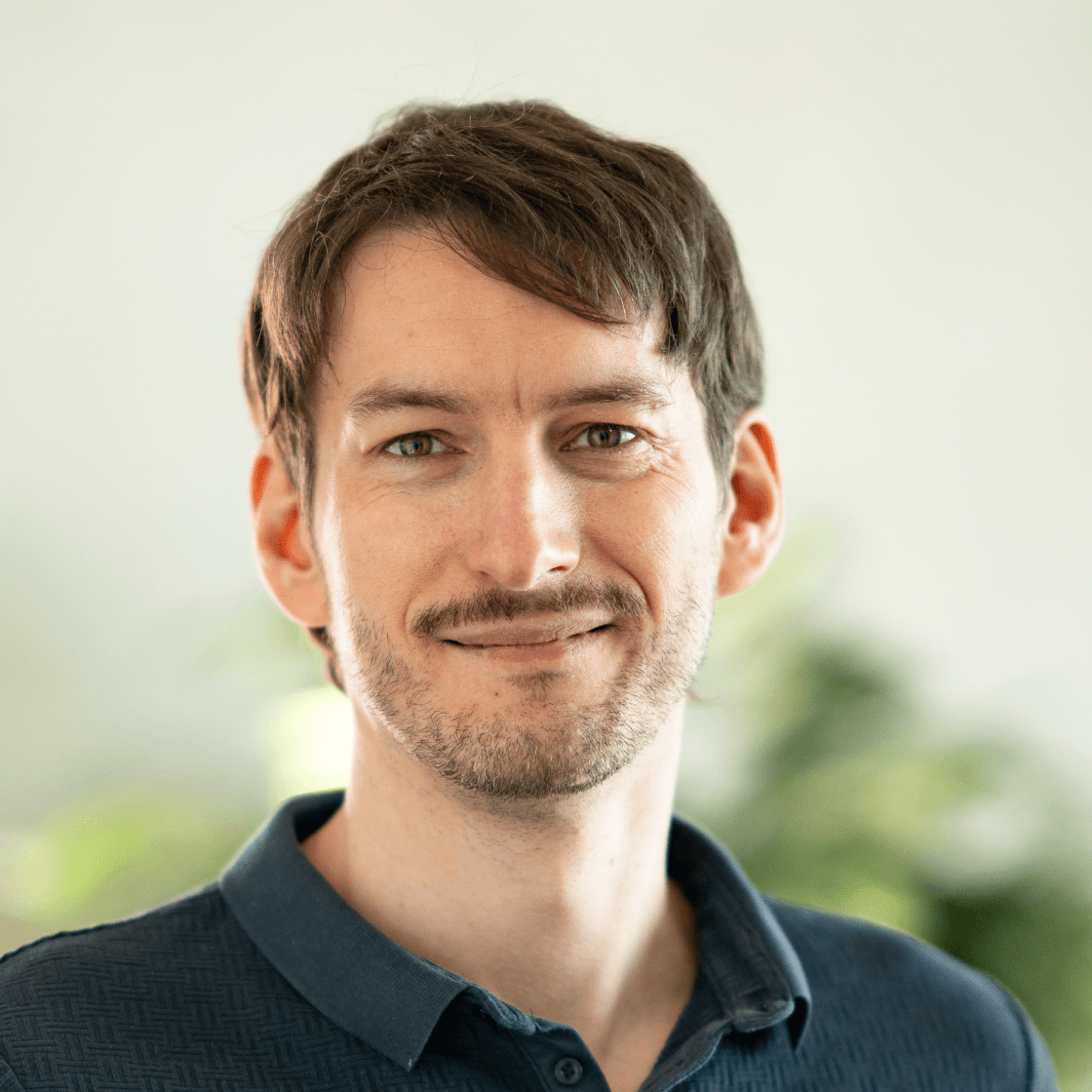 Porträt von Peter mit braunen Haaren, kurzem Bart und dunkelblauem Poloshirt. Er blickt freundlich und kompetent in die Kamera. Im Hintergrund sorgen unscharfe Pflanzen für eine natürliche, helle und freundliche Atmosphäre – typisch für moderne Büro- oder Homeoffice-Umgebungen.