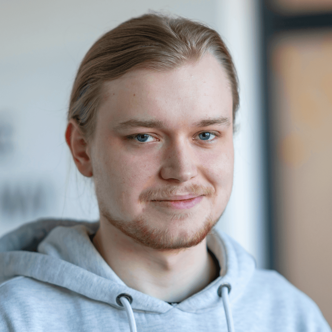Porträt von Alexander mit langen, hellblonden Haaren, die zu einem Zopf gebunden sind. Er trägt einen hellgrauen Hoodie und lächelt dezent in die Kamera. Der Hintergrund ist unscharf und zeigt eine moderne Indoor-Umgebung in hellen Farben.