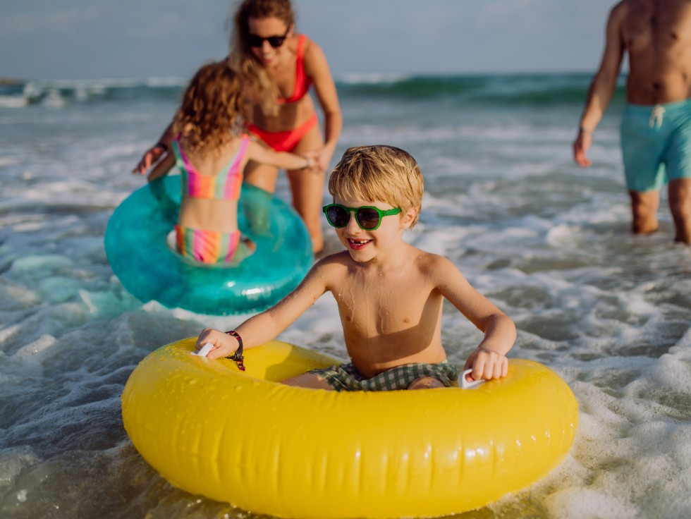 Kinder spielen mit Schwimmreifen im Meer, zwei Erwachsene im Hintergrund