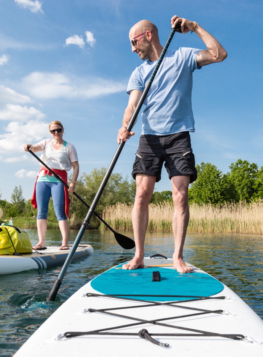Zwei Menschen stehend auf Stand-Up-Paddle-Boards auf dem Wasser, das Ufer gesäumt mit Gräsern