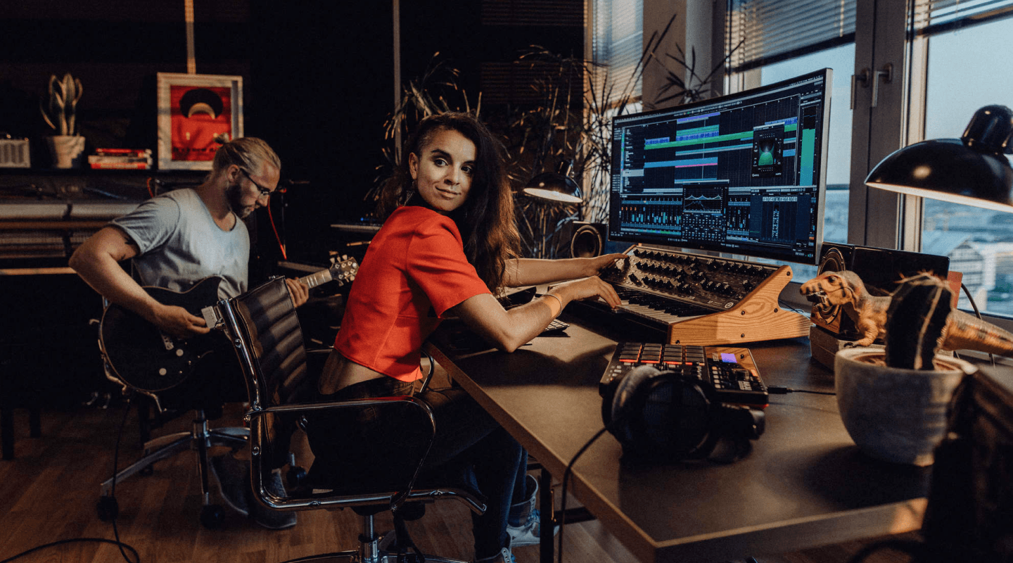 Ein Mann spielt Gitarre im sitzen. Eine Frau sitzt daneben und stellt an Reglern von Tontechnik. Vor den Beiden ist ein großer Monitor auf dem die Steinberg Software zu sehen ist.