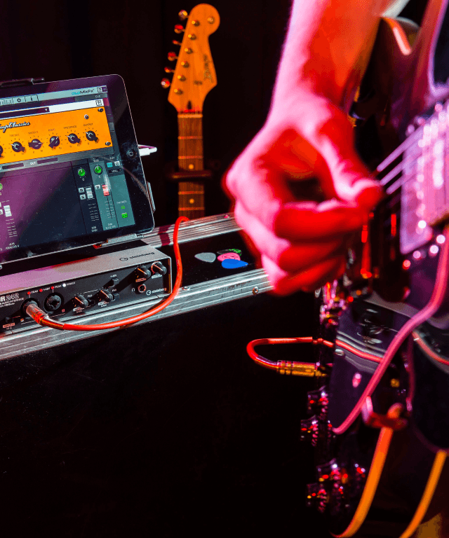 Detailaufnahme von einer Hand, die Gitarre spielt. Im Hintergrund Tontechnik.