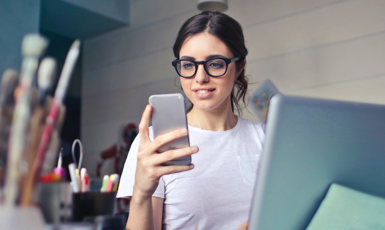 Frau sitzt am Labtop und schaut auf ihr Smartphone, was sie in der Hand hält. Auf dem Tisch stehen noch Pinsel und Stifte.
