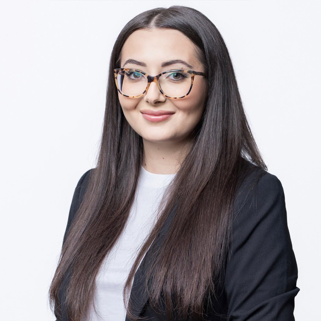 Mia Angelov trägt ihr langes, glattes, dunkelbraunes Haare offen und eine große braune Brille. Sie lächelt leicht in die Kamera.