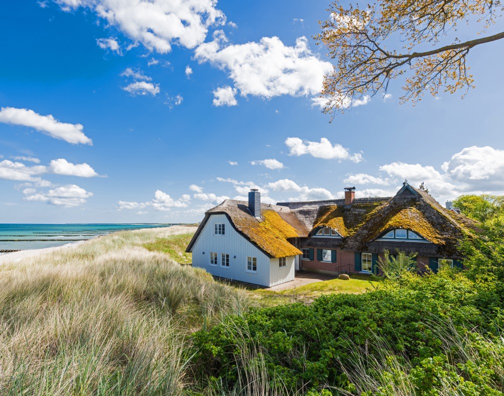 Ein großes, reetgedecktes Bauernshaus steht direkt in den Dünen. Dahinter ist das Meer zu sehen.