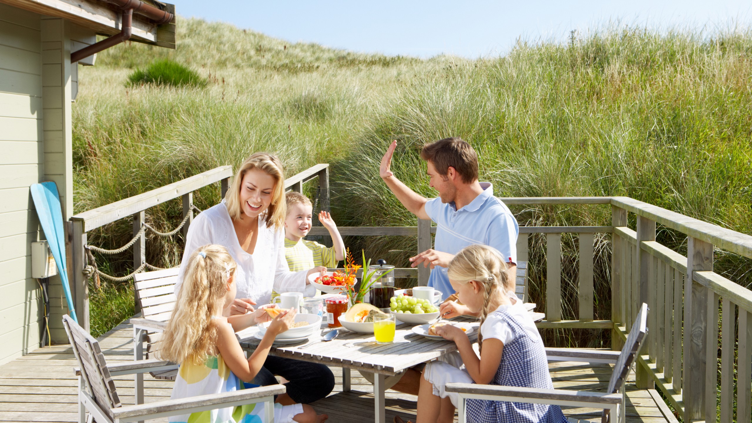 Eine Familie mit Mutter, Vater und zwei Töchtern und einem Sohn sitzen an einem Frühstückstisch auf einer Holzterasse in den Dünen.