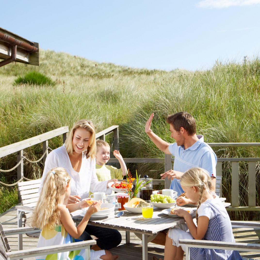 Eine Familie mit Mutter, Vater und zwei Töchtern und einem Sohn sitzen an einem Frühstückstisch auf einer Holzterasse in den Dünen.