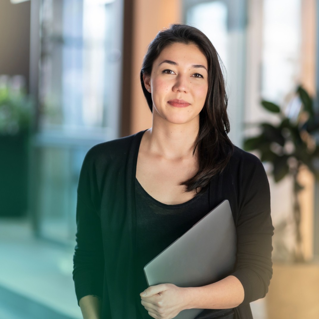 Eine selbstbewusste junge Frau steht in einem hellen BĂŒrokorridor und hĂ€lt ein geschlossenes Laptop in einer Hand.