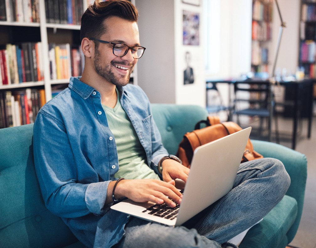 Foto: Junger Mann mit kurzen, braunen Haaren und eckiger Brille sitzt im Schneidersitz auf einer blauen Couch, hat einen aufgeklappten Laptop auf dem Schoß, bllickt lächelnd auf den Bildschirm und tippt auf der Tastatur.