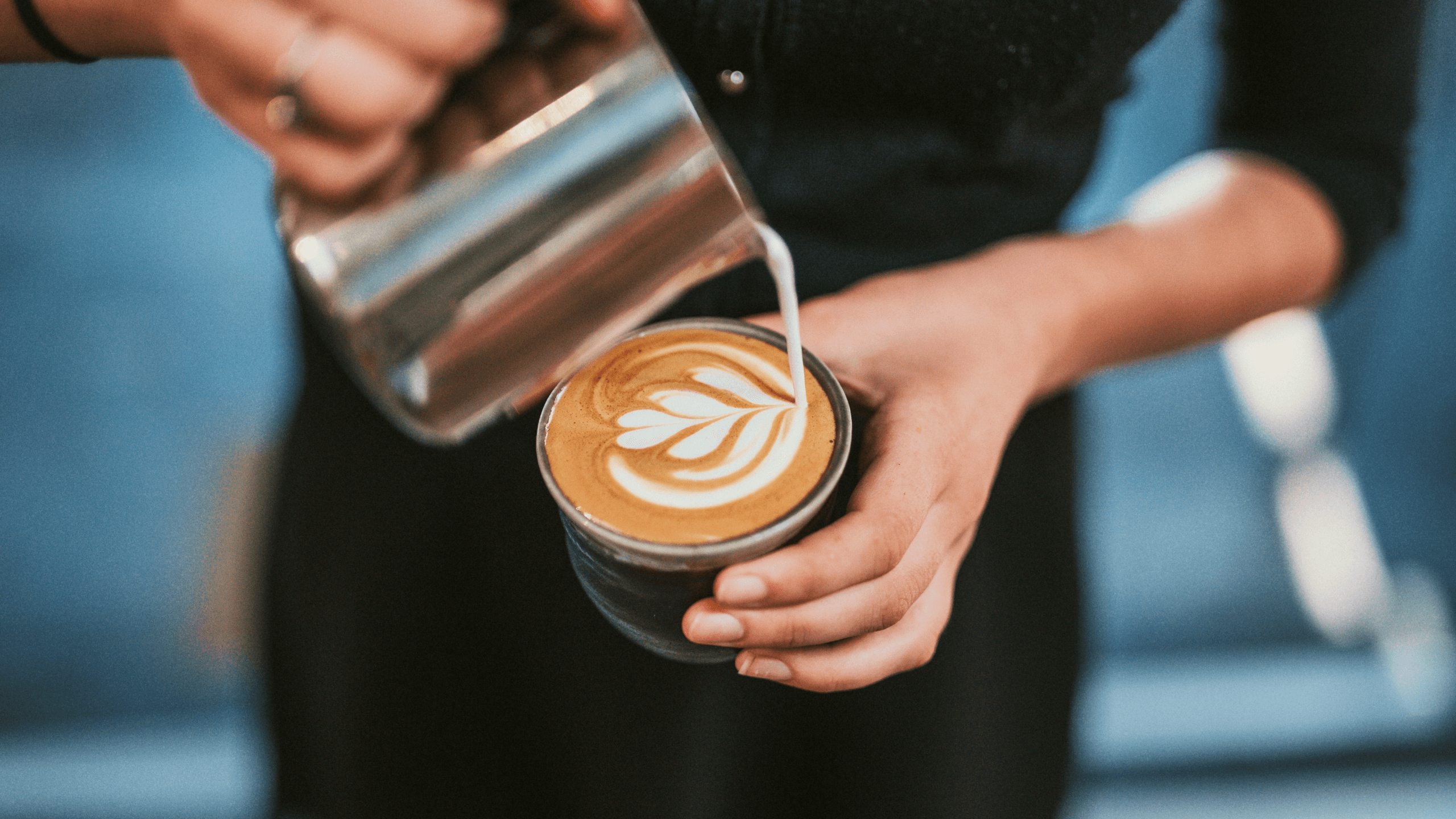 Foto: Nahaufnahme der Hände und des Oberkörpers einer Person, die Milchschaum kunstvoll in eine Kaffeetasse eingießt
