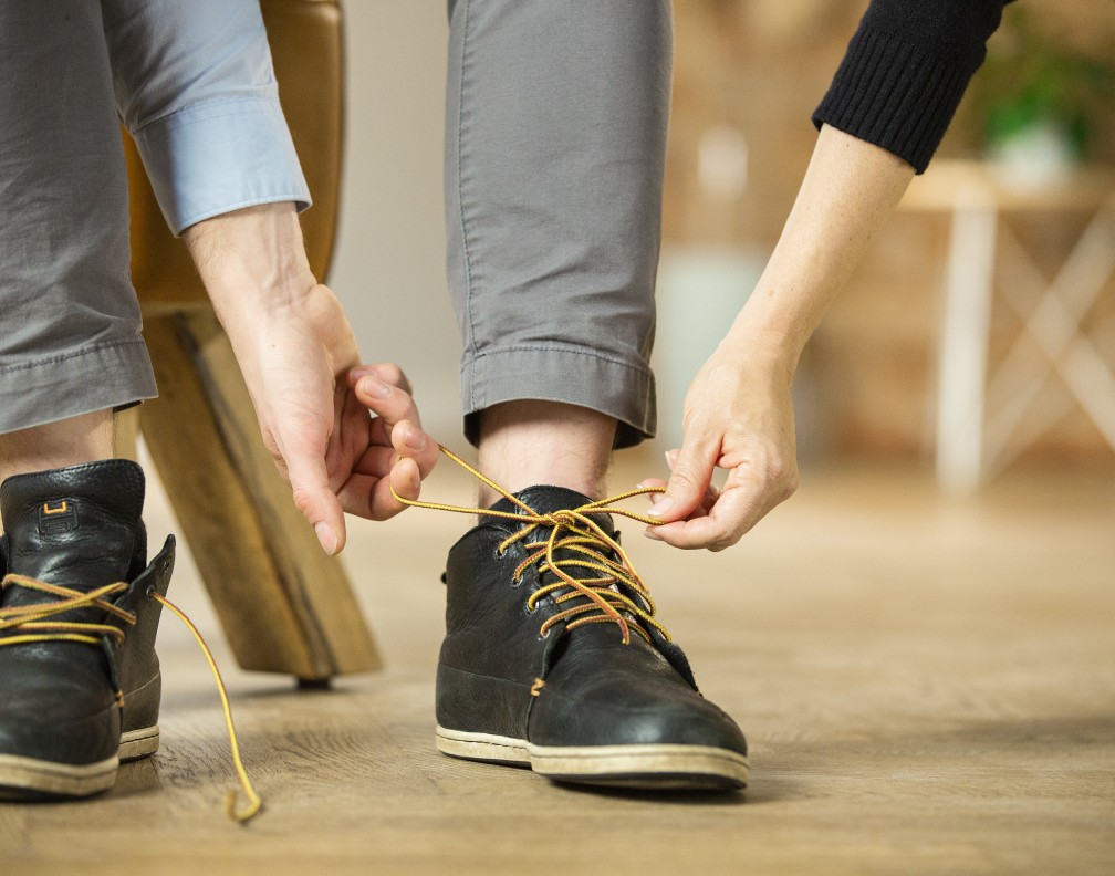 Schwarze Herren-Schuhe mit gelben Schnürsenkeln. Eine männliche und eine weibliche Hand greifen nach der gebundenen Schleife am Schuh.