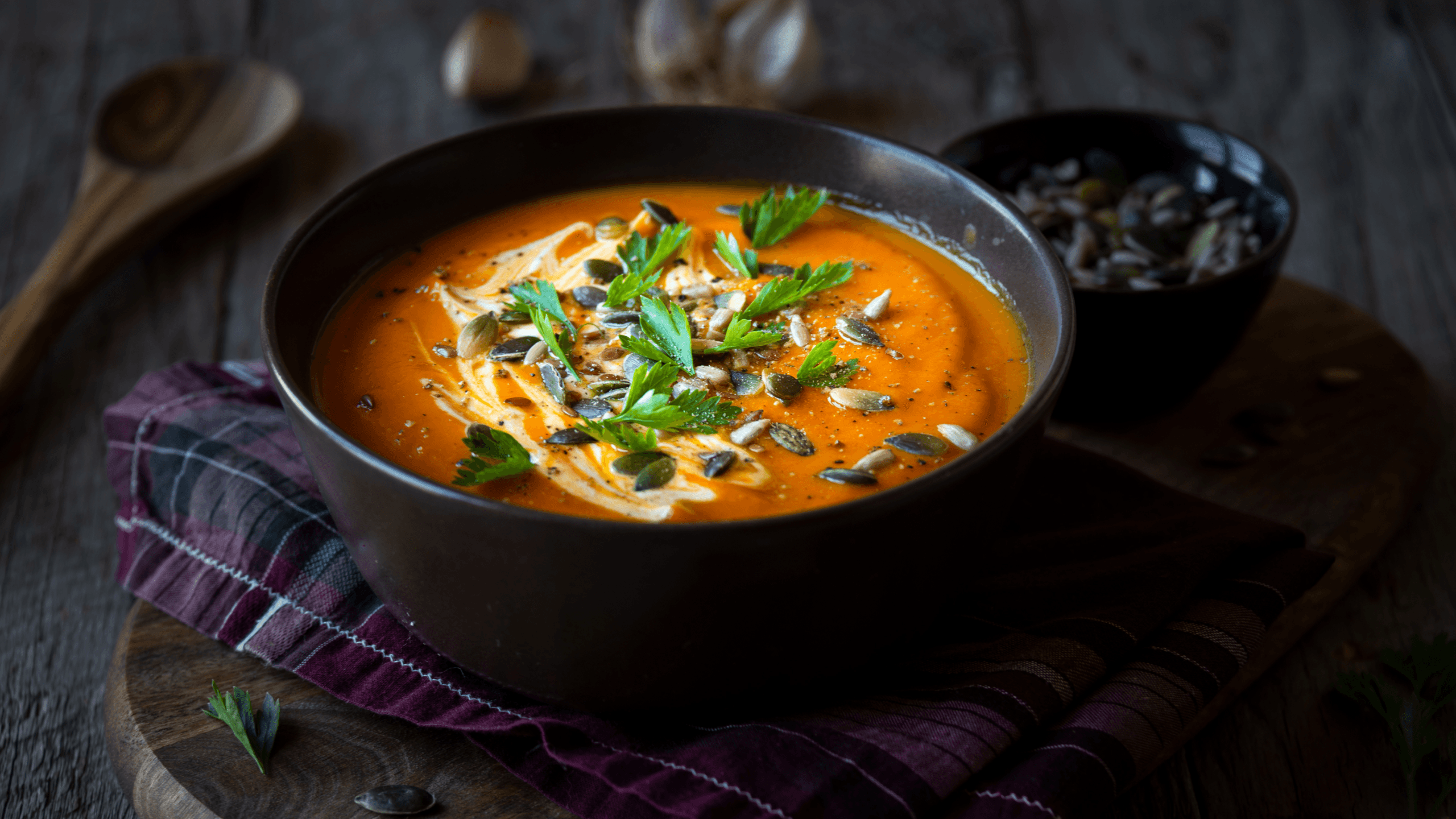 Eine Schüssel cremige Kürbissuppe, kunstvoll garniert mit Kürbiskernen, frischen Kräutern und einem Sahne-Topping. Die Schüssel steht auf einem rustikalen Holztisch, dekoriert mit einem dunklen Tuch. Im Hintergrund ist ein Holzlöffel sichtbar. Kulinarische Exzellenz der Gourmetfabrik.