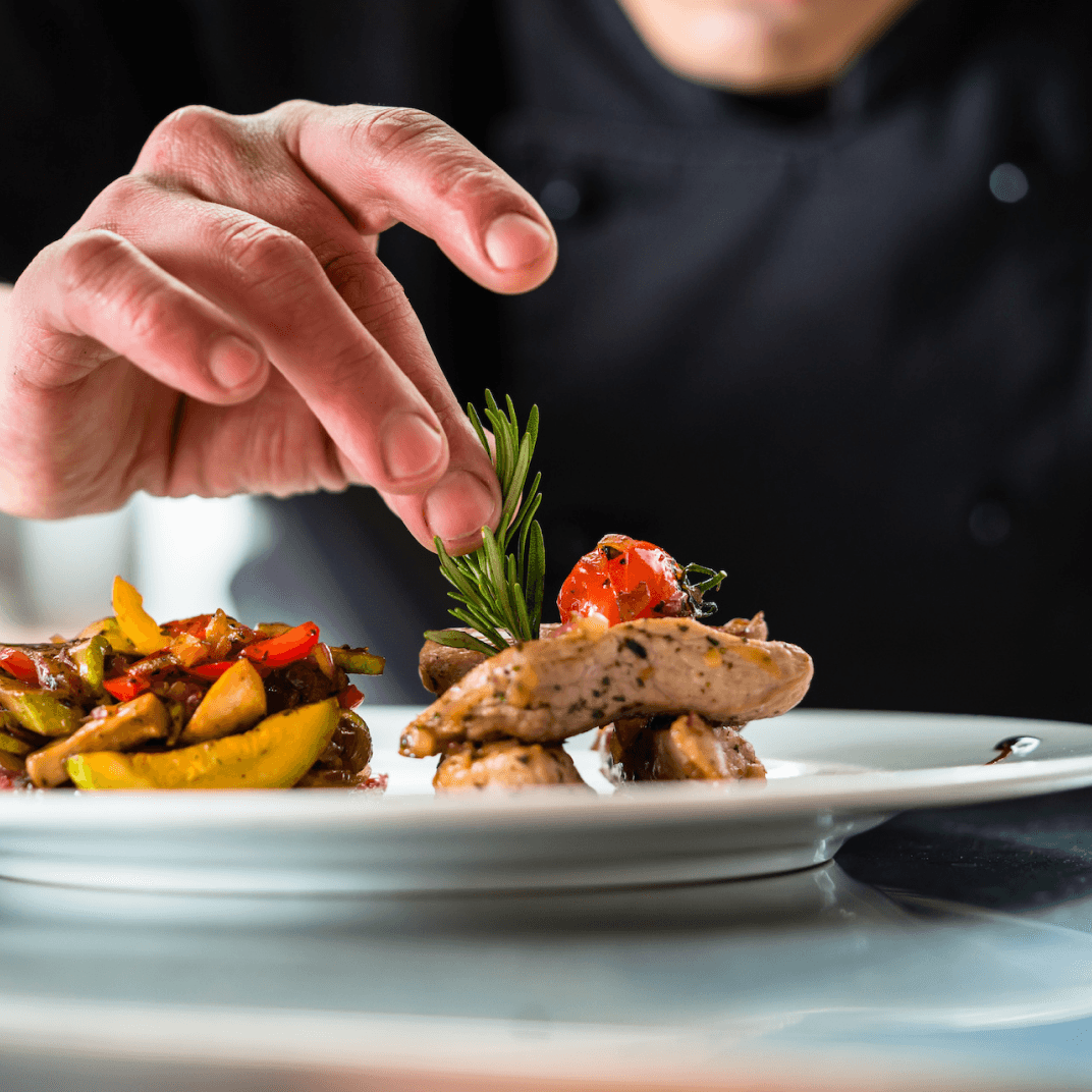 Ein Koch in schwarzer Uniform arrangiert sorgfältig ein Gourmetgericht auf einem weißen Teller. Das Gericht besteht aus zartem Fleisch, verziert mit einem Rosmarinzweig, und einer bunten Beilage aus sautiertem Gemüse. Kulinarische Perfektion in der Gourmetfabrik.