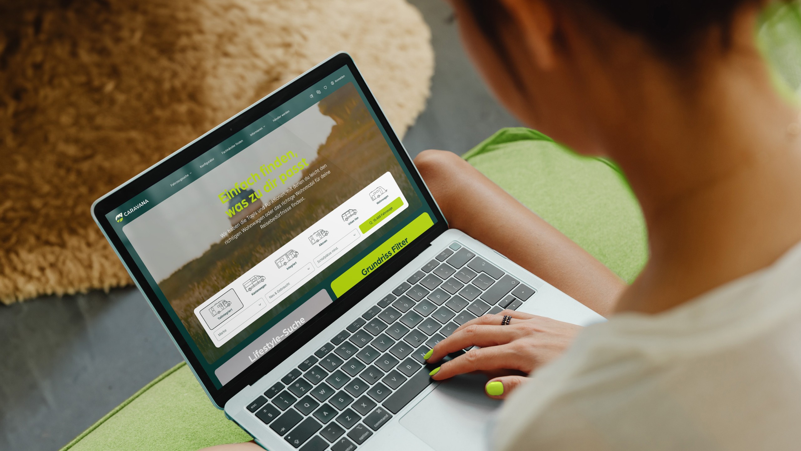 Frau sitzt auf einem grünen Sofa und nutzt einen Laptop, um auf der CARAVANA-Website nach verschiedenen Wohnmobiltypen zu suchen. Der Bildschirm zeigt Optionen zur Auswahl von Teilintegriert, Alkoven, Vollintegriert und Kastenwagen, sowie einen Grundriss Filter. Die Frau hat grün lackierte Fingernägel und trägt einen Ring.