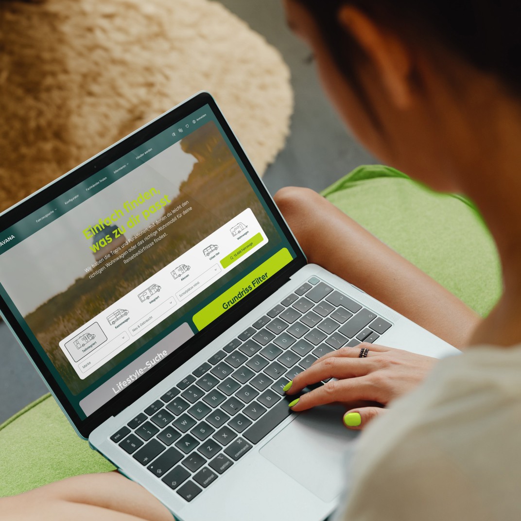Frau sitzt auf einem grünen Sofa und nutzt einen Laptop, um auf der CARAVANA-Website nach verschiedenen Wohnmobiltypen zu suchen. Der Bildschirm zeigt Optionen zur Auswahl von Teilintegriert, Alkoven, Vollintegriert und Kastenwagen, sowie einen Grundriss Filter. Die Frau hat grün lackierte Fingernägel und trägt einen Ring.