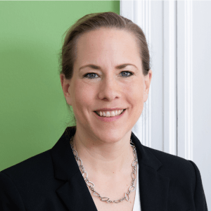 Portrait von Dr. Katharina Reuter, Geschäftsführerin des Bundesverbands Nachhaltige Wirtschaft (BNW) e.V. Sie trägt ein schwarzes Blazer und ein silbernes Kettchen. Dr. Reuter hat braune Haare, blaue Augen und lächelt in die Kamera. Der Hintergrund ist hellgrün und weiß.