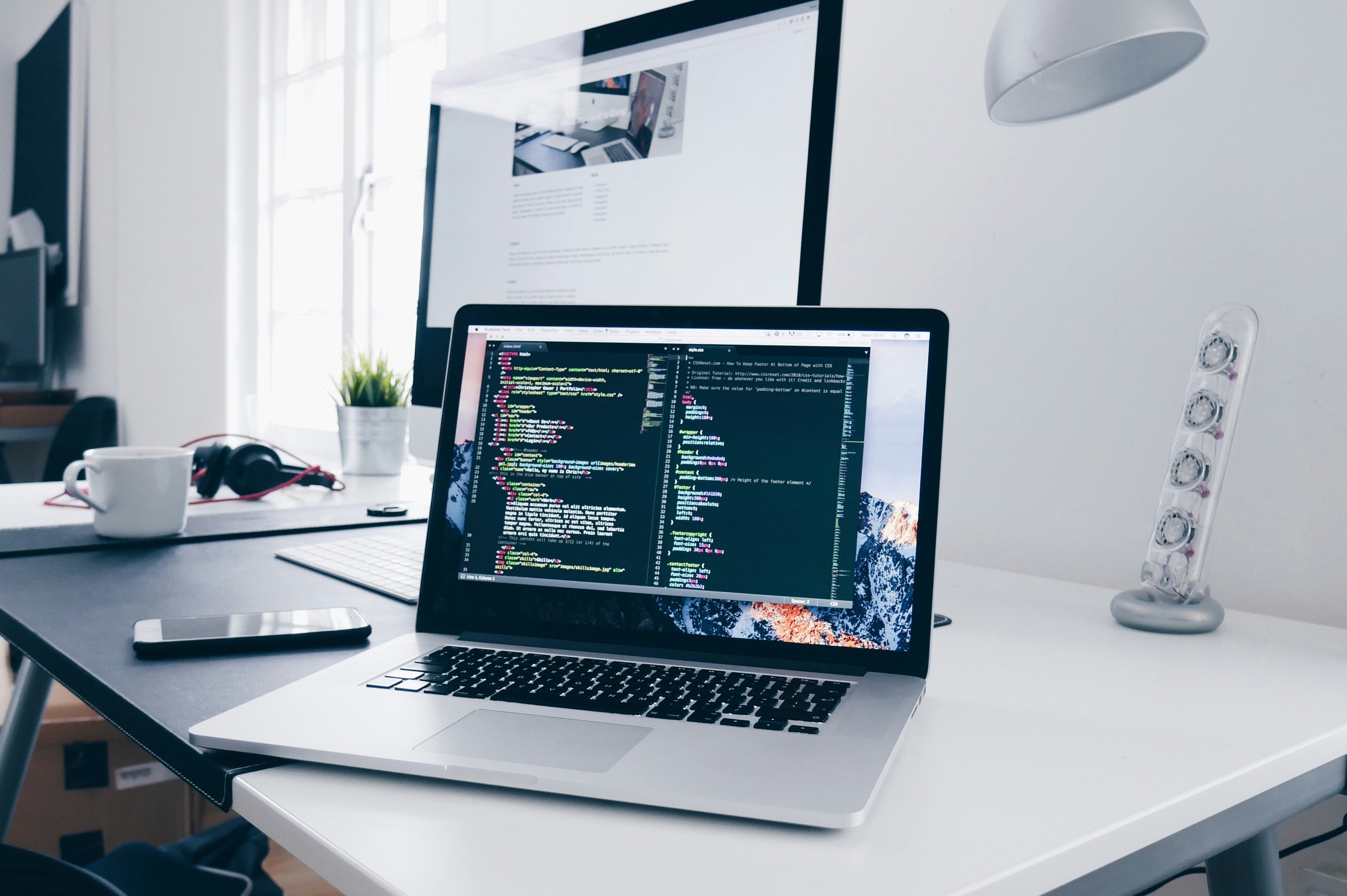 Laptop auf einem Schreibtisch mit geöffnetem Code-Editor, im Hintergrund ein großer Monitor und verschiedene Büromaterialien.