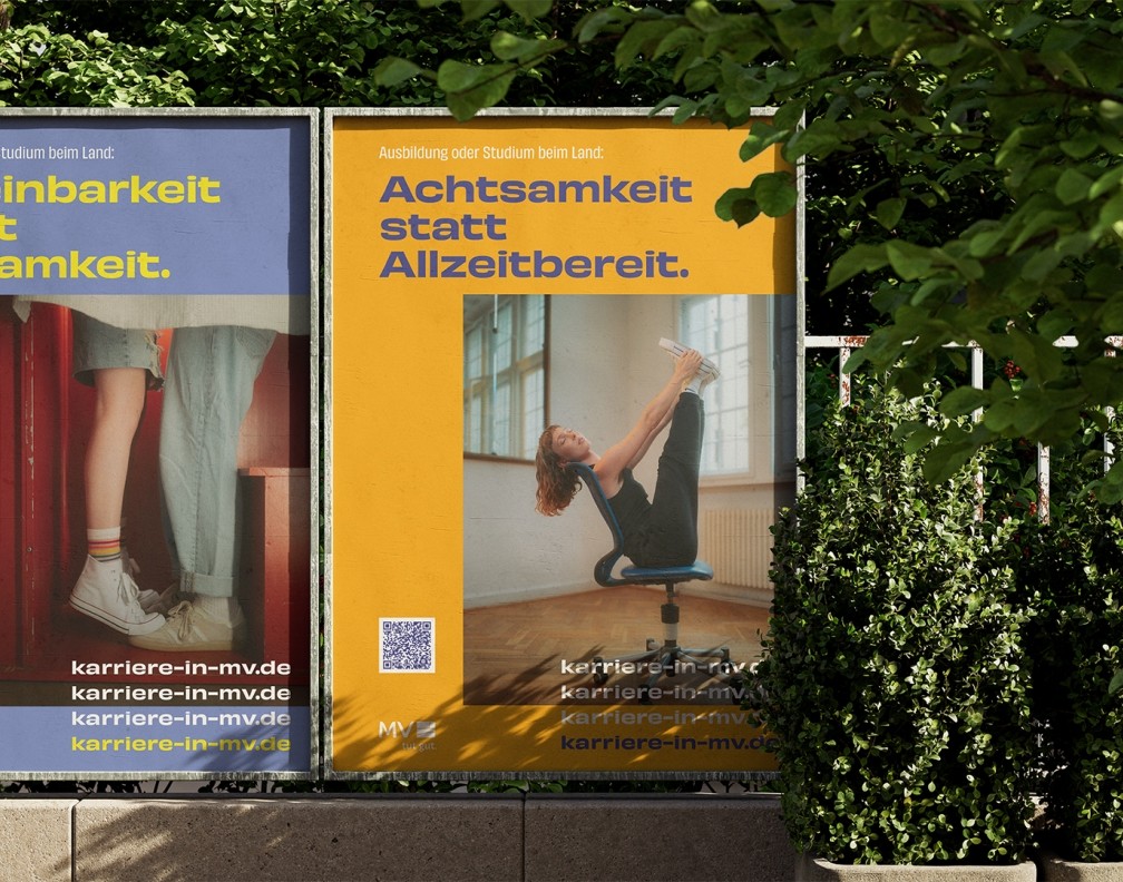 Zwei Werbeplakate im Freien mit den Slogans „Achtsamkeit statt Allzeitbereit“ und „Einbarkeit statt Einsamkeit“ für Karrieren beim Land. Das linke Plakat zeigt zwei Personen, die in Turnschuhen auf einer roten Treppe stehen. Das rechte Plakat zeigt eine Frau, die beide Beine auf einem Bürostuhl in die Höhe streckt. Beide Plakate enthalten die Webadresse „karriere-in-mv.de“ und einen QR-Code für weitere Informationen. Die Plakate sind von grüner Vegetation umgeben.
