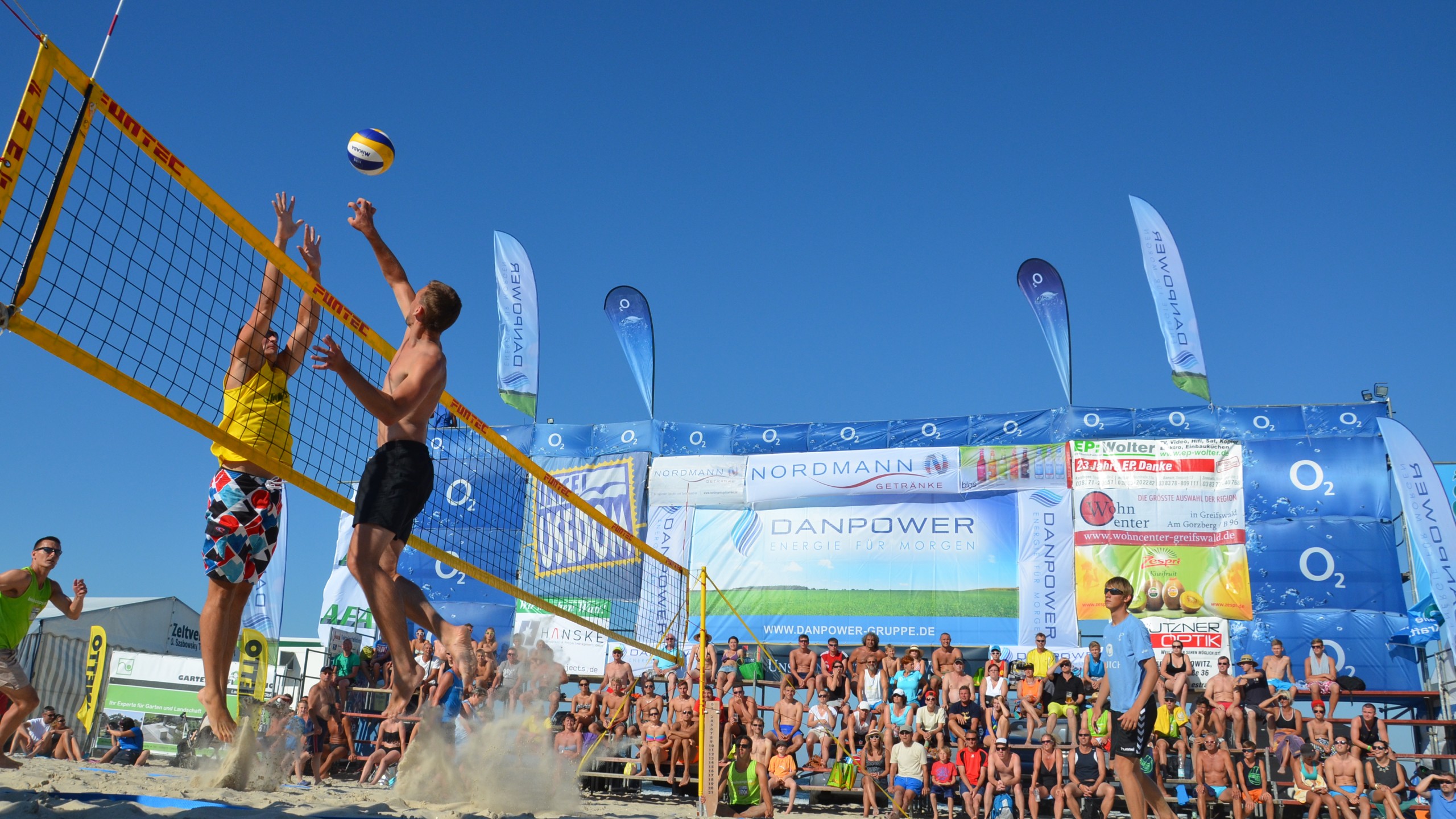 Spannendes Beachvolleyball-Match am Strand der Insel Usedom bei strahlend blauem Himmel. Zwei Spieler im Sprung am Netz, umgeben von jubelnden Zuschauern auf Tribünen. Im Hintergrund sind Sponsorenbanner und Strandatmosphäre sichtbar. Perfekte Darstellung von Sport und Freizeit am Strand.