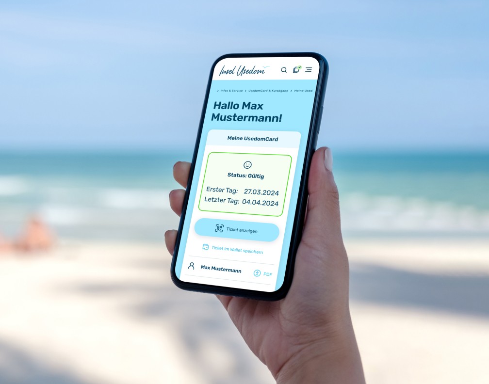 Eine Person hält ein Smartphone am Strand, auf dem die digitale 'UsedomCard' angezeigt wird. Der Bildschirm zeigt persönliche Informationen wie Status 'Gültig', Erster und Letzter Tag sowie Optionen wie 'Ticket anzeigen' und 'Im Wallet speichern'. Im Hintergrund das Meer und ein Sandstrand, was eine moderne und benutzerfreundliche Urlaubserfahrung vermittelt.