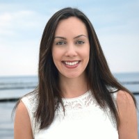 Porträt einer lächelnden Frau mit dunklem Haar und weißem Oberteil vor einer Küstenlandschaft mit blauem Himmel und Meer im Hintergrund, ideal für professionelle oder persönliche Präsentationen.