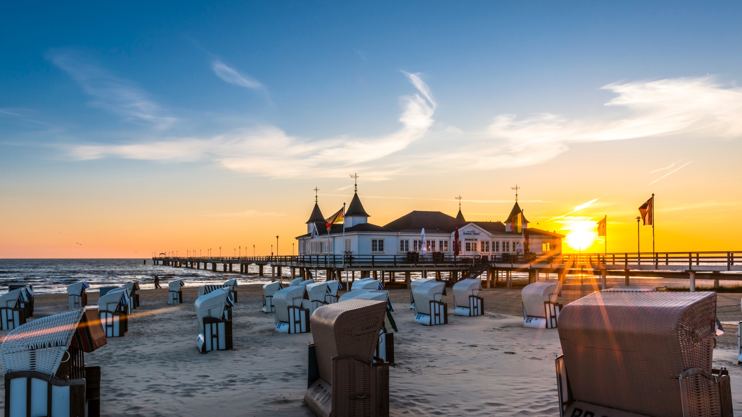 Abendliche Strandansicht der Seebrücke Heringsdorf auf der Insel Usedom bei Sonnenuntergang. Strandkörbe im Vordergrund und die Seebrücke mit ihrem historischen Bauwerk in der Mitte, eingerahmt von der leuchtenden Sonne und einem klaren Himmel. Perfekte Kulisse für einen romantischen Ostseeurlaub.