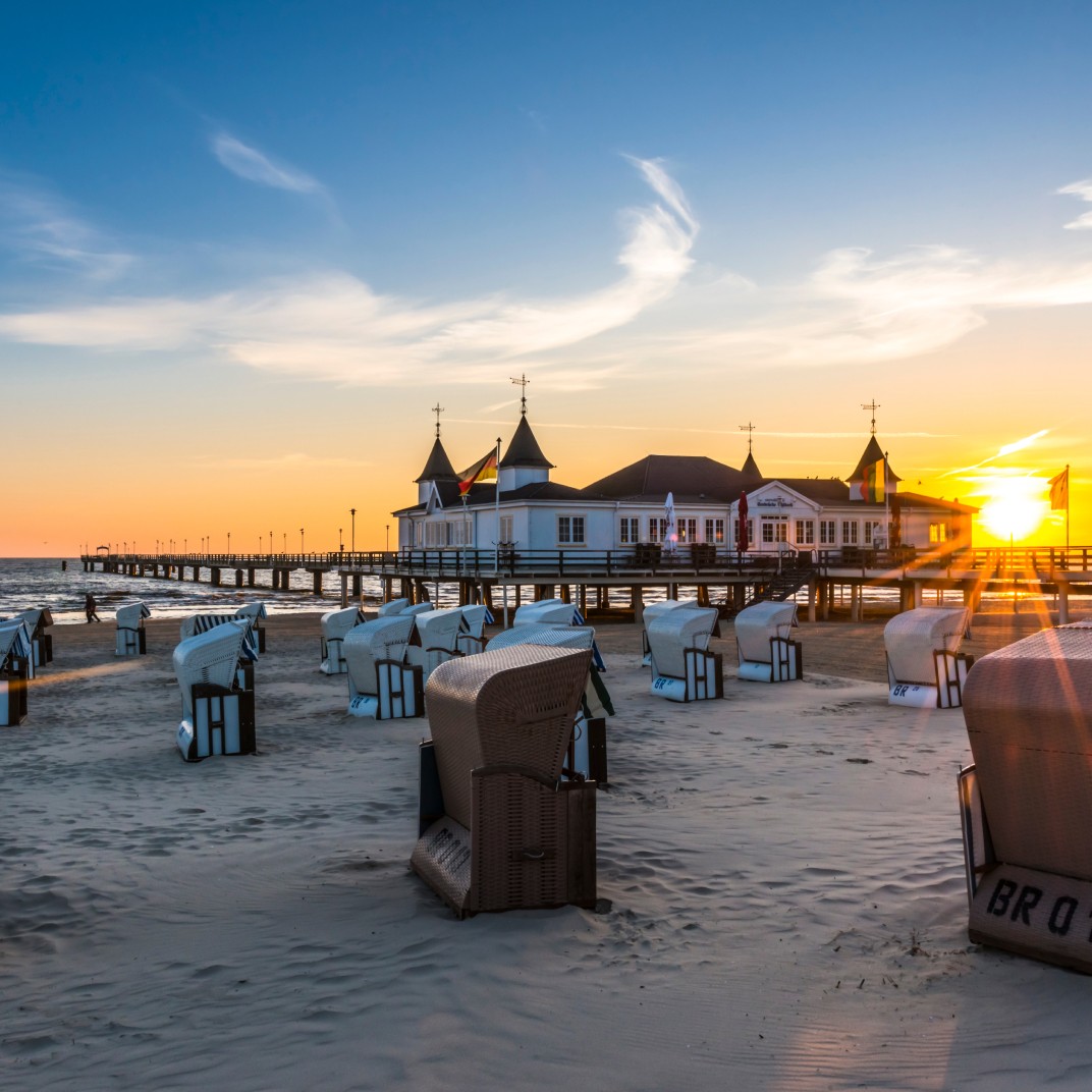 Abendliche Strandansicht der Seebrücke Heringsdorf auf der Insel Usedom bei Sonnenuntergang. Strandkörbe im Vordergrund und die Seebrücke mit ihrem historischen Bauwerk in der Mitte, eingerahmt von der leuchtenden Sonne und einem klaren Himmel. Perfekte Kulisse für einen romantischen Ostseeurlaub.