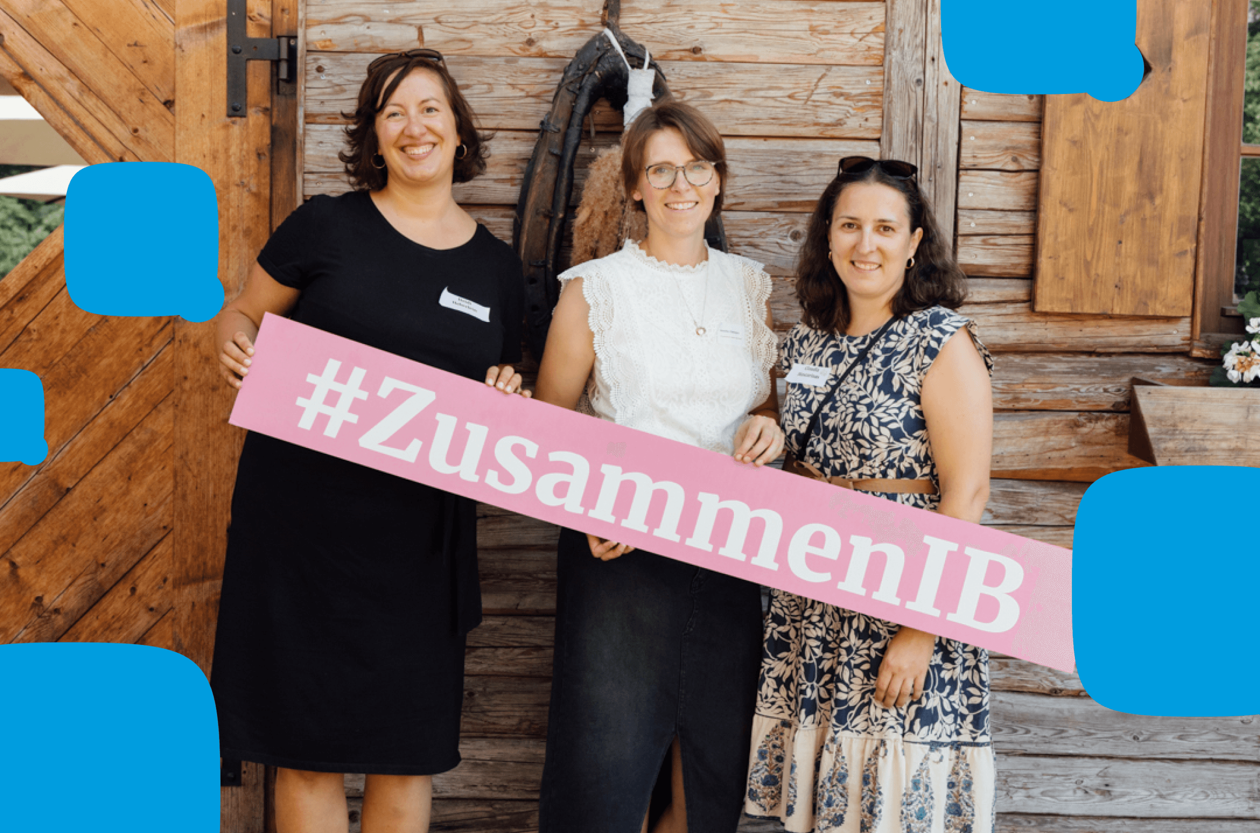 Drei lächelnde Frauen aus dem Team des Internationalen Bunds (IB) stehen vor einer rustikalen Holzwand und halten ein rosa Schild mit der Aufschrift „#ZusammenIB“. Die Szene strahlt Gemeinschaft und Teamgeist aus.