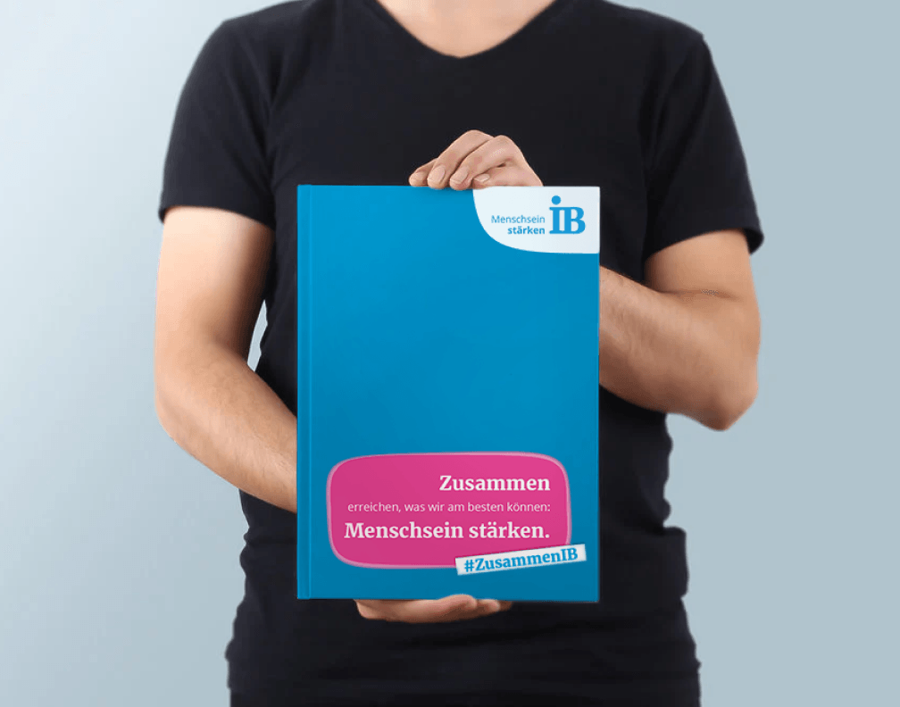 Eine Person in schwarzem T-Shirt hält ein blaues Plakat mit dem Logo des Internationalen Bunds (IB) und dem Slogan „Zusammen erreichen, was wir am besten können: Menschsein stärken.“ sowie dem Hashtag #ZusammenIB. Der Hintergrund ist neutral in hellem Grau gehalten.