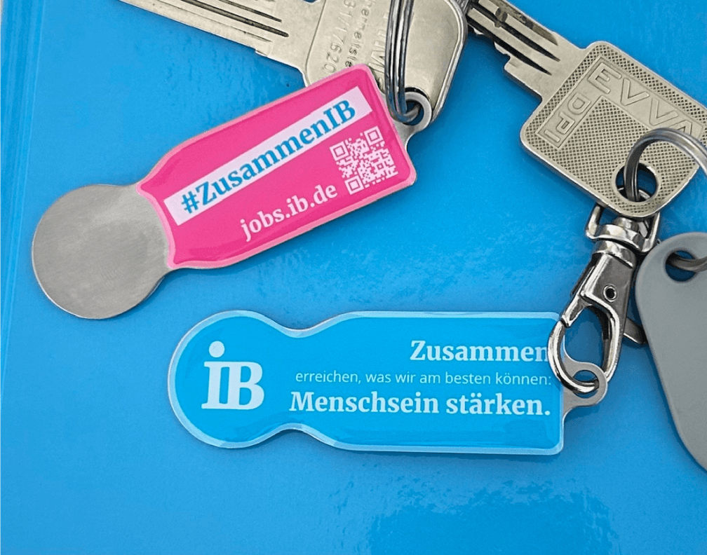 Ein Schlüsselbund mit zwei Einkaufswagenchips des Internationalen Bunds (IB) liegt auf einer blauen Oberfläche. Der pinke Chip zeigt den Hashtag „#ZusammenIB“ und die Website „jobs.ib.de“ mit einem QR-Code. Der blaue Chip trägt den Slogan „Zusammen erreichen, was wir am besten können: Menschsein stärken.“.