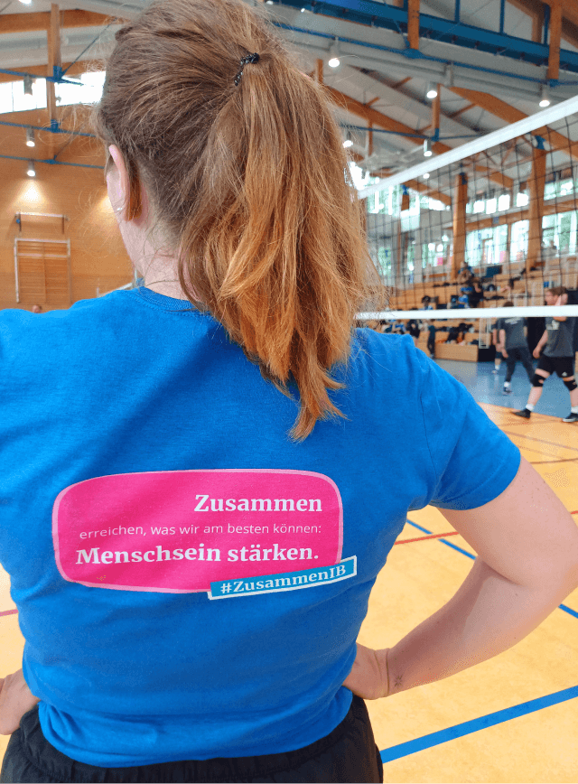 Rückansicht einer Frau in einem blauen T-Shirt mit dem Aufdruck „Zusammen erreichen, was wir am besten können: Menschsein stärken.“ und dem Hashtag #ZusammenIB. Sie steht in einer Sporthalle, im Hintergrund sind Volleyballspieler und eine Tribüne zu sehen.
