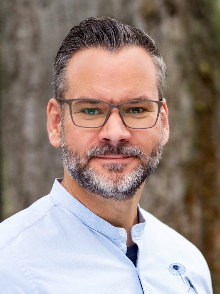 Christof lächelt und trägt eine Brille, graumeliertem Bart und nach hinten gestyltem Haar, in einem hellblauen Hemd vor einem natürlichen Hintergrund mit Bäumen.