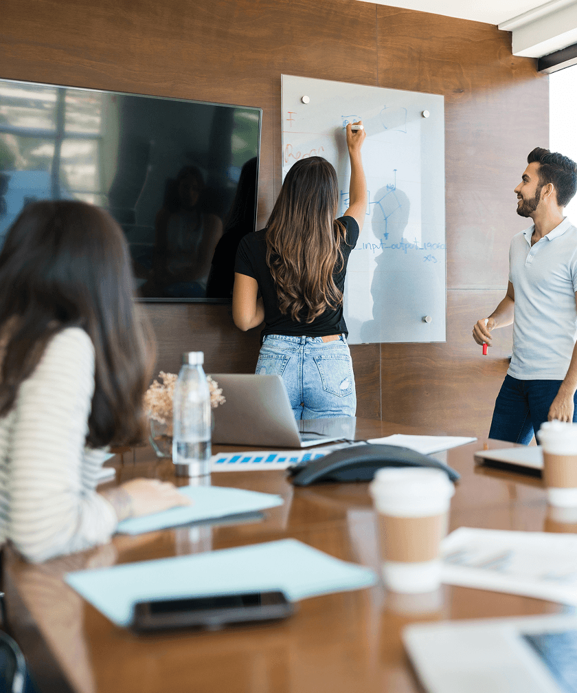 Team-Meeting in modernem Büro: Eine Frau schreibt auf eine transparente Tafel, während Kollegen aufmerksam zuhören. Die Szene zeigt eine professionelle Arbeitsumgebung mit Laptops, Notizen, Kaffee und einem großen Bildschirm. Zusammenarbeit, Brainstorming und kreative Ideenfindung im Business-Kontext.