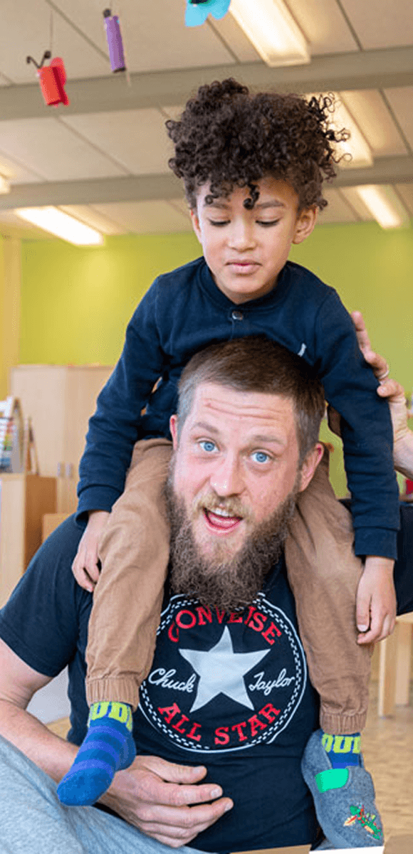 Männliche Fachkraft des Internationalen Bundes mit fröhlichem Kind auf den Schultern – spielerische Betreuung und starke Bindung in der Kindertagesstätte.