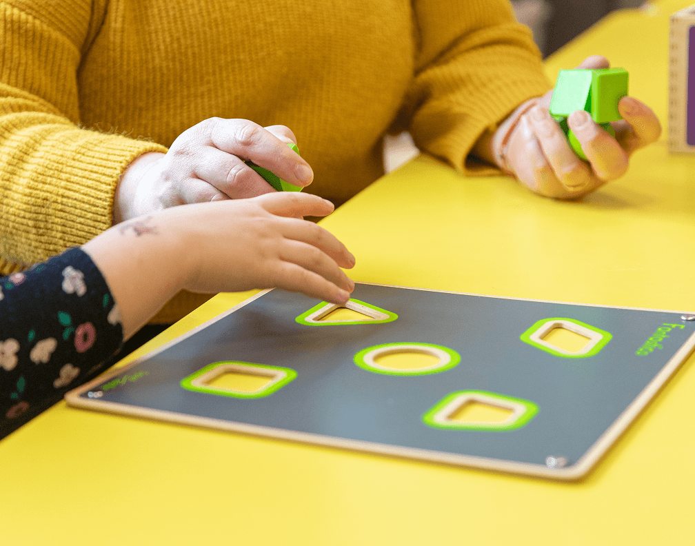 Frühkindliche Förderung beim Internationalen Bund – pädagogische Fachkraft unterstützt Kind beim Lernen mit einem Formensortierspiel zur Entwicklung motorischer und kognitiver Fähigkeiten.