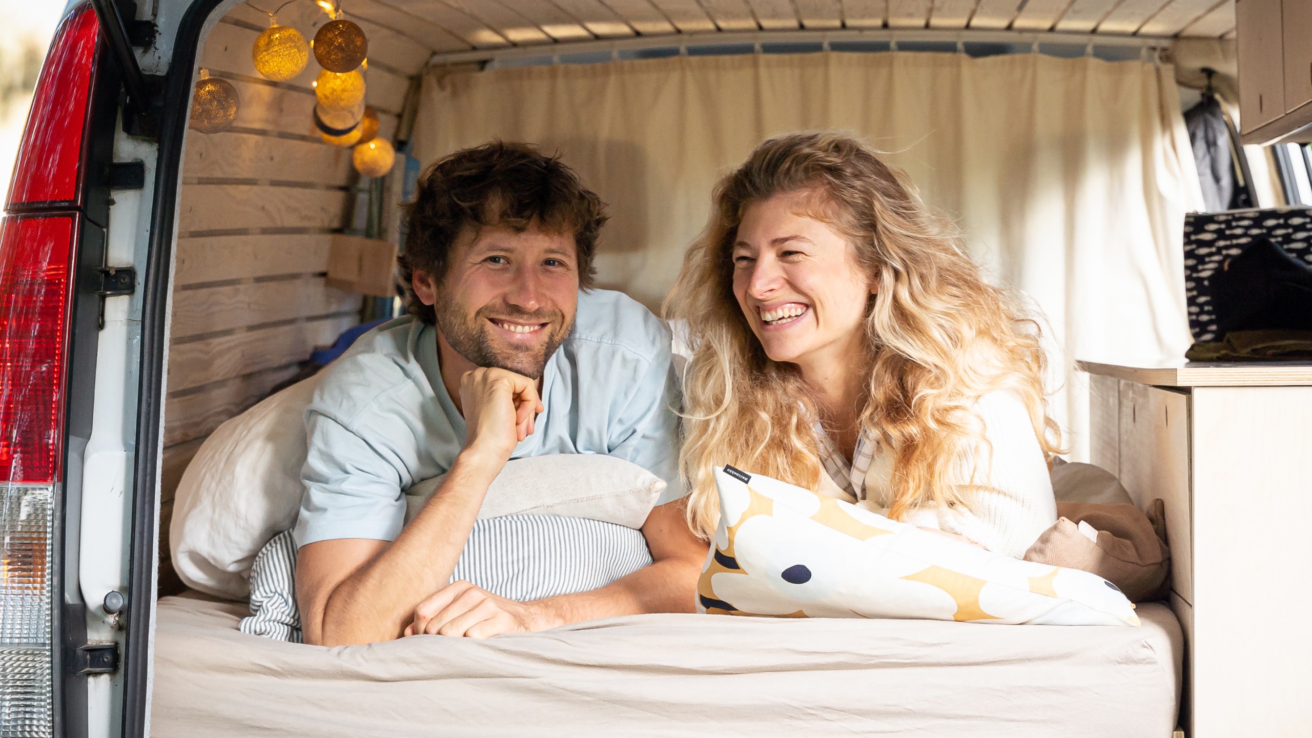 Ein Mann und eine Frau liegen in einem ausgebauten Campervan, schauen lächelnd in die Kamera