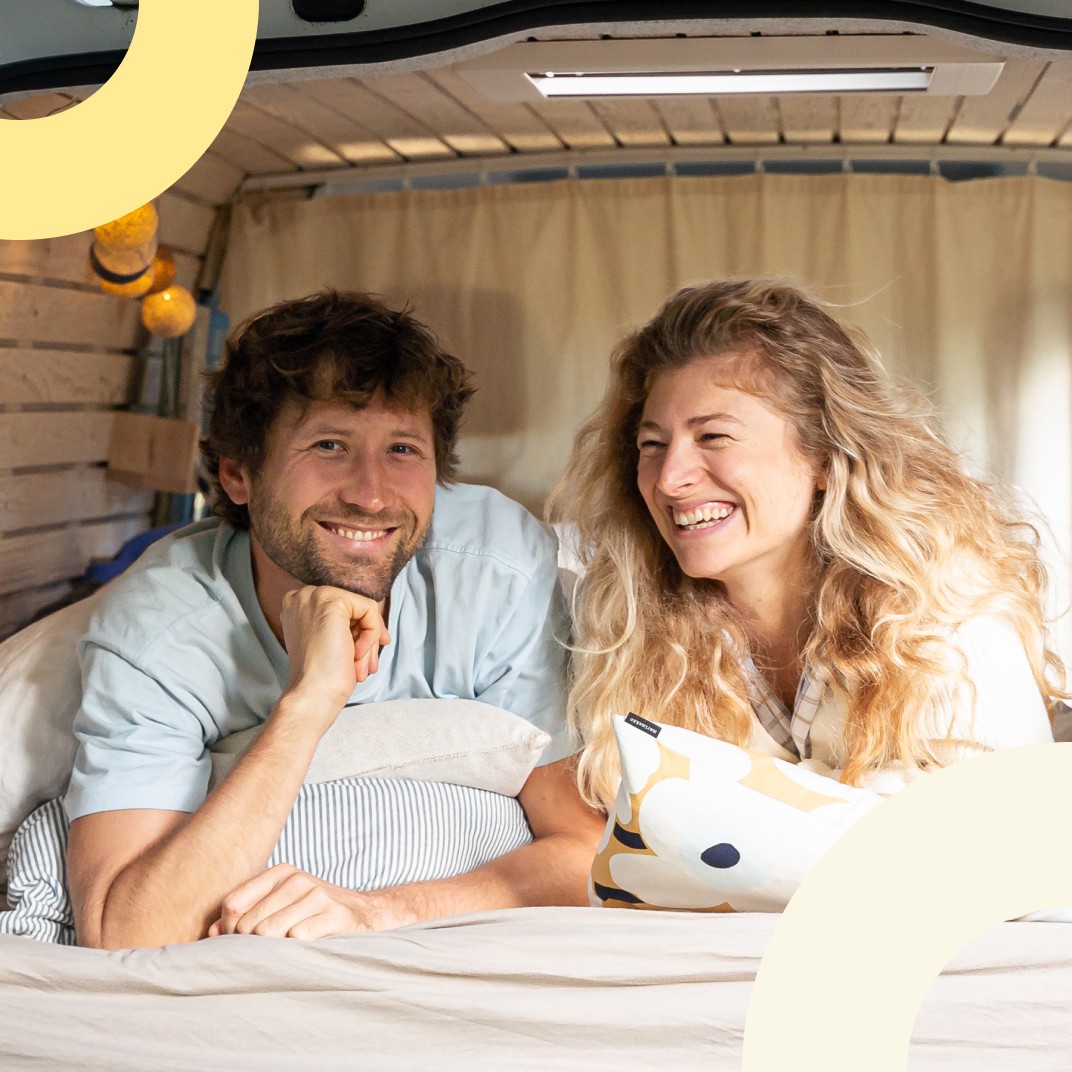 Ein Mann und eine Frau liegen in einem ausgebauten Campervan, schauen lächelnd in die Kamera