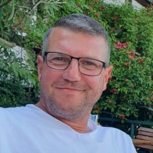 Mann mit grauer Kurzhaarfrisur, eckiger schwarzer Brille, weißem T-Shirt vor grünem Busch im Hintergrund