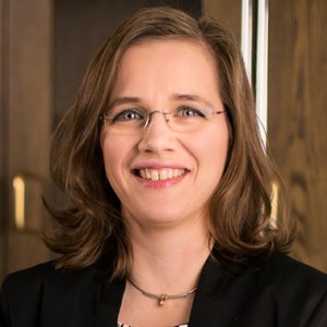 Frau mit braunen mittellangen Haaren, randloser Brille, schwarzem Blazer, vor einem Schrank aus Holz