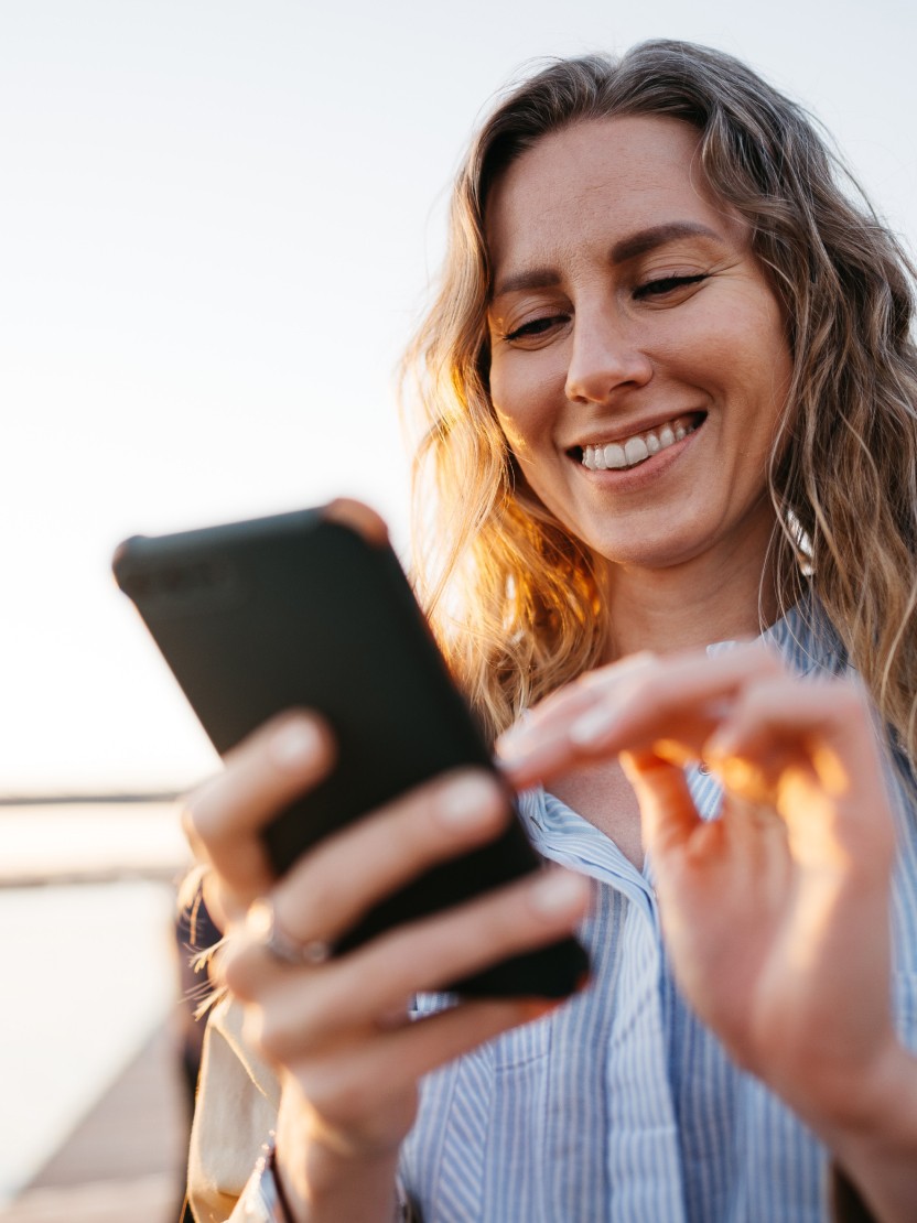 Eine Frau tippt glücklich auf ihrem Handy, im Hintergrund ist verschwommen Wasser zu erkennen