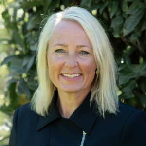 Frau mit blonen Haaren und schwarzem Oberteil vor grünem Busch im Hintergrund