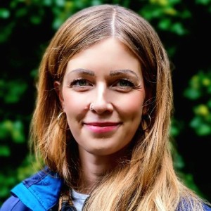 Frau mit hellblonden Haaren, blauer Jacke vor grünem Busch im Hintergrund