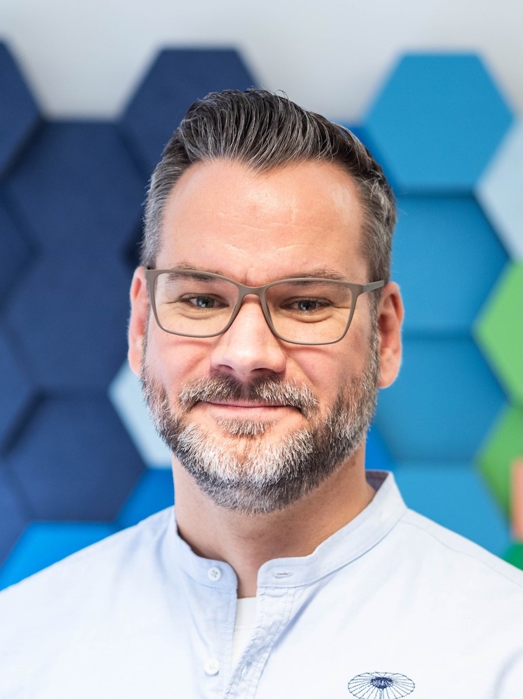 Christof lächelt und trägt eine Brille, graumeliertem Bart und nach hinten gestyltem Haar, in einem hellblauen Hemd vor einem Hintergrund mit sechseckigen Schallschutzkachen.
