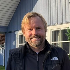 Hauke Schürmann steht mit schwarzer Jacke von einem hellblauen Blockhaus und lacht