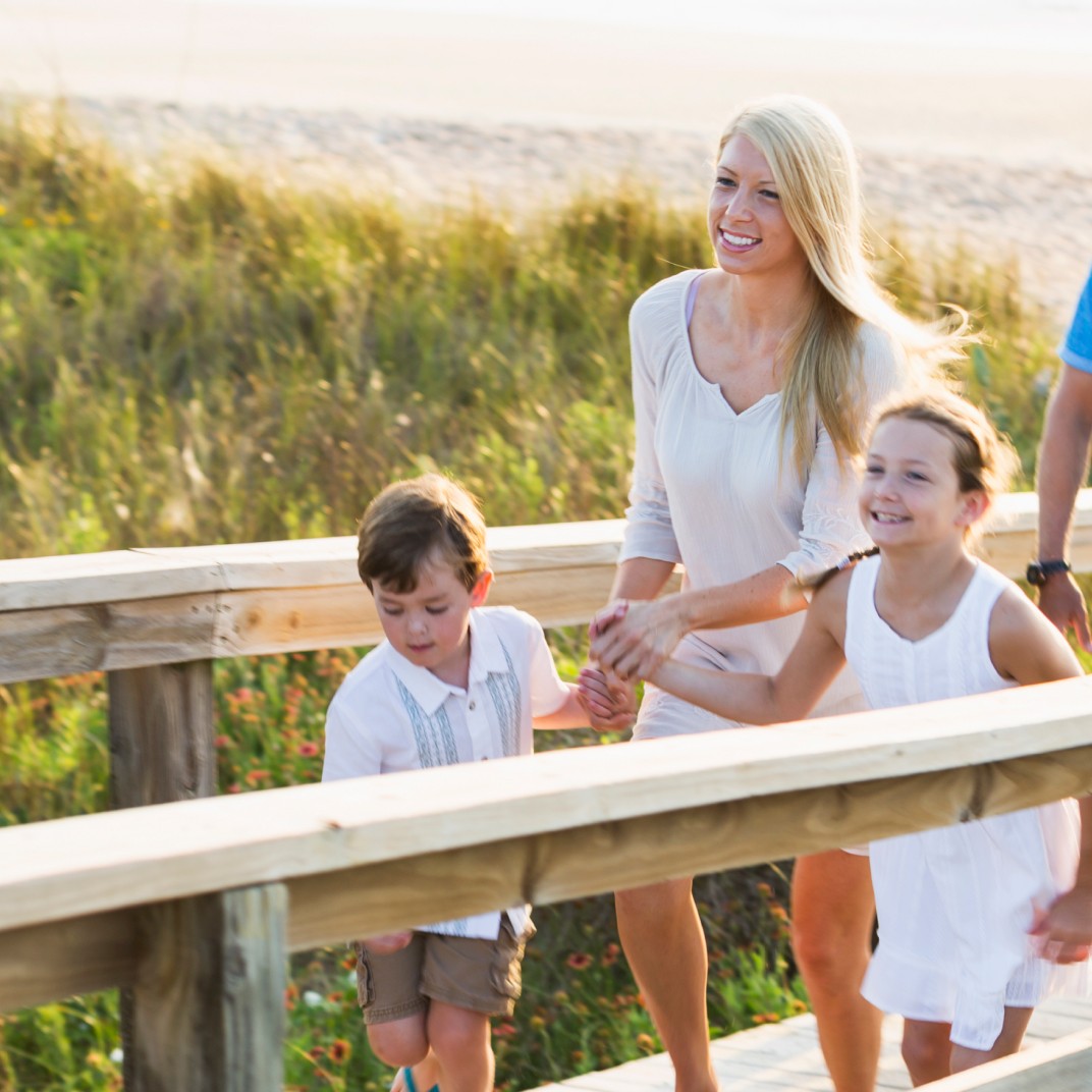 Zwei Erwachsene und zwei Kinder laufen einen Steg hinauf, im Hintergrund Strand und Meer