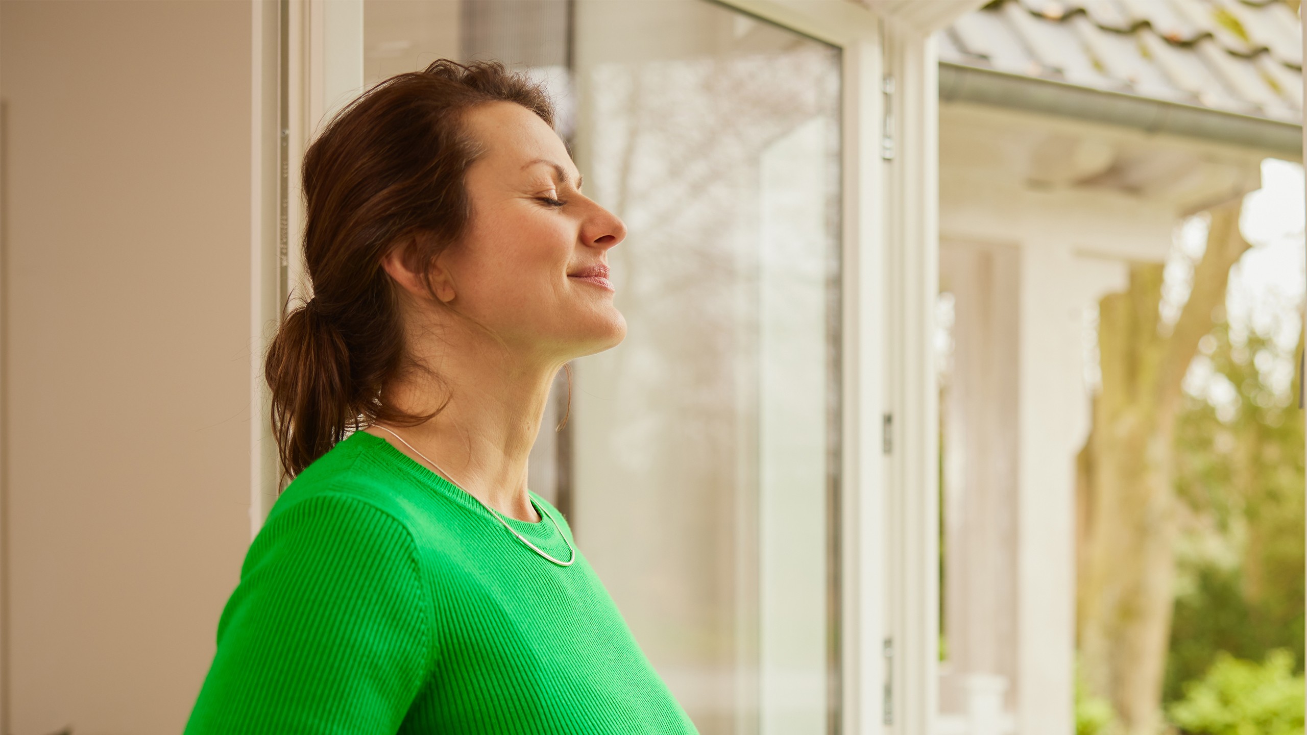 Frau mit grünem Oberteil steht mit geschlossenen Augen vor geöffnetem Fenster