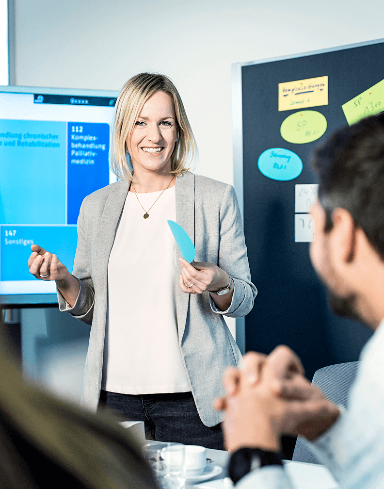 Team-Meeting in modernem Büro: Eine Frau hält Notizzettel, während Kollegen aufmerksam zuhören. Die Szene zeigt eine professionelle Arbeitsumgebung mit Notizen, Kaffee und einem großen Bildschirm. Zusammenarbeit, Brainstorming und kreative Ideenfindung im Business-Kontext.