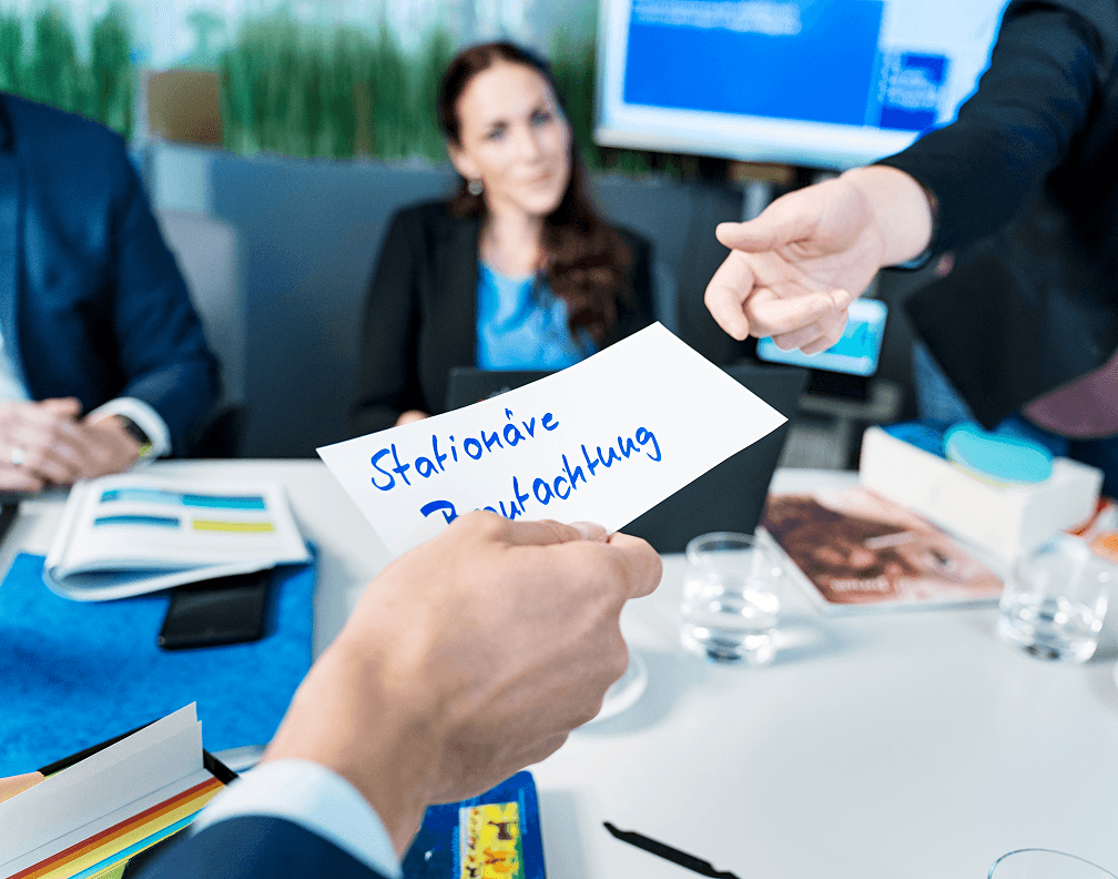 Nahaufnahme eines weißen Notizzettels, auf dem handschriftlich "Stationäre Begutachtung" steht. Der Zettel wird während eines Meetings an Kolleg:innen weitergereicht.